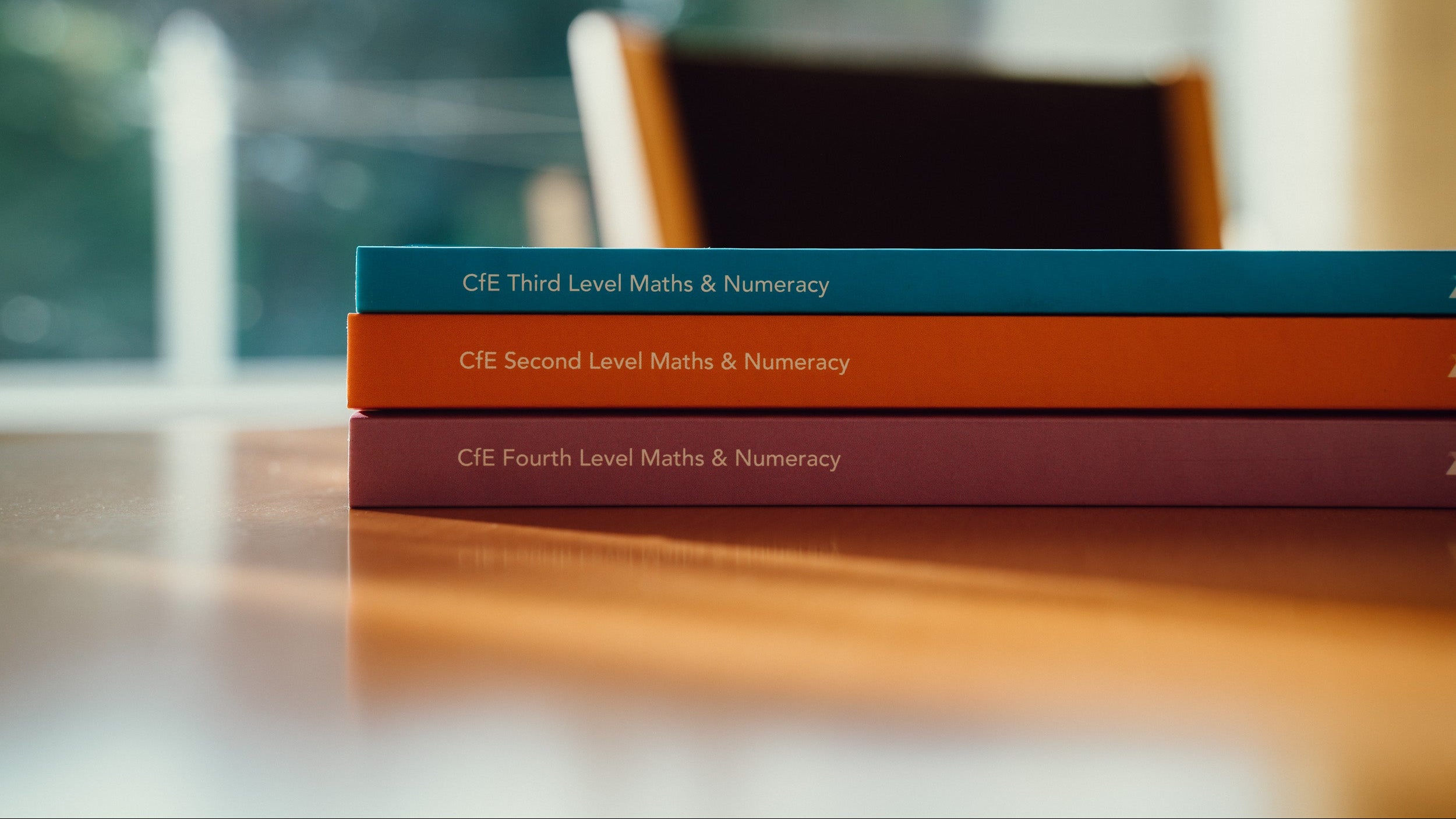 Three books titled 'CIE Third Level Maths & Numeracy', 'CIE Second Level Maths & Numeracy', and 'CIE Fourth Level Maths & Numeracy' on a wooden surface.