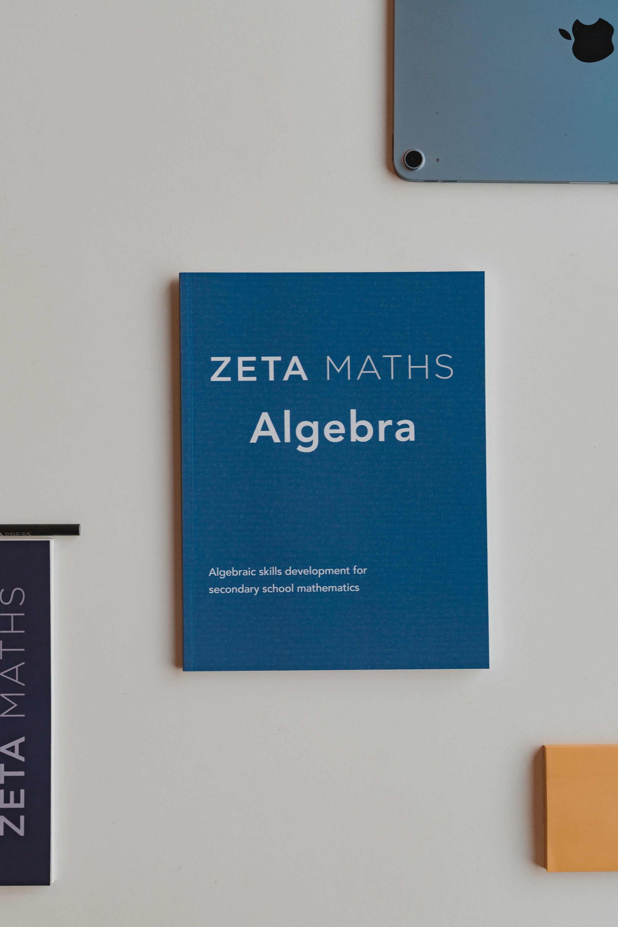 Blue book titled 'Zeta Maths Algebra' on a light gray surface with a laptop and other books in the background.